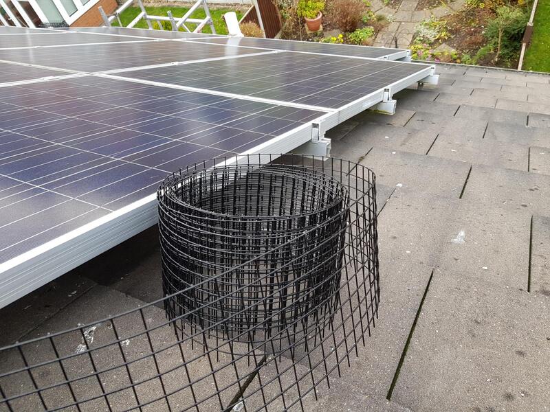 A roll of black bird-proofing wire mesh sitting on a tiled roof beside a solar panel array.