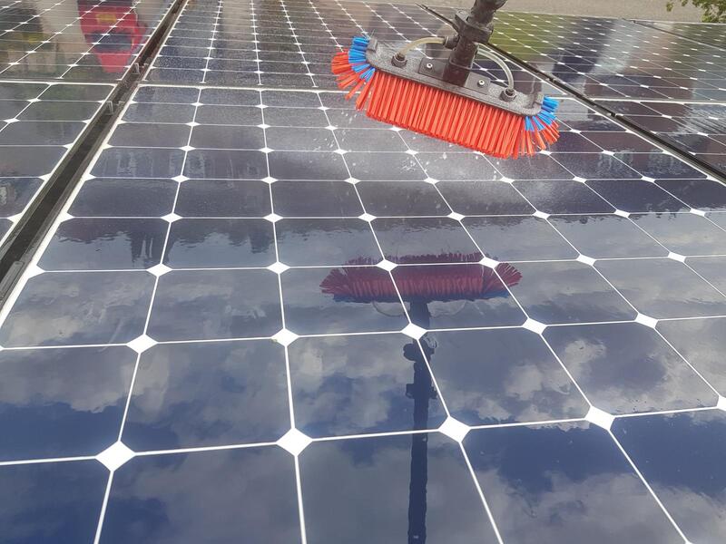 A row of solar panels being washed with a pole-mounted brush, water and cloud reflections visible on the glossy surface.